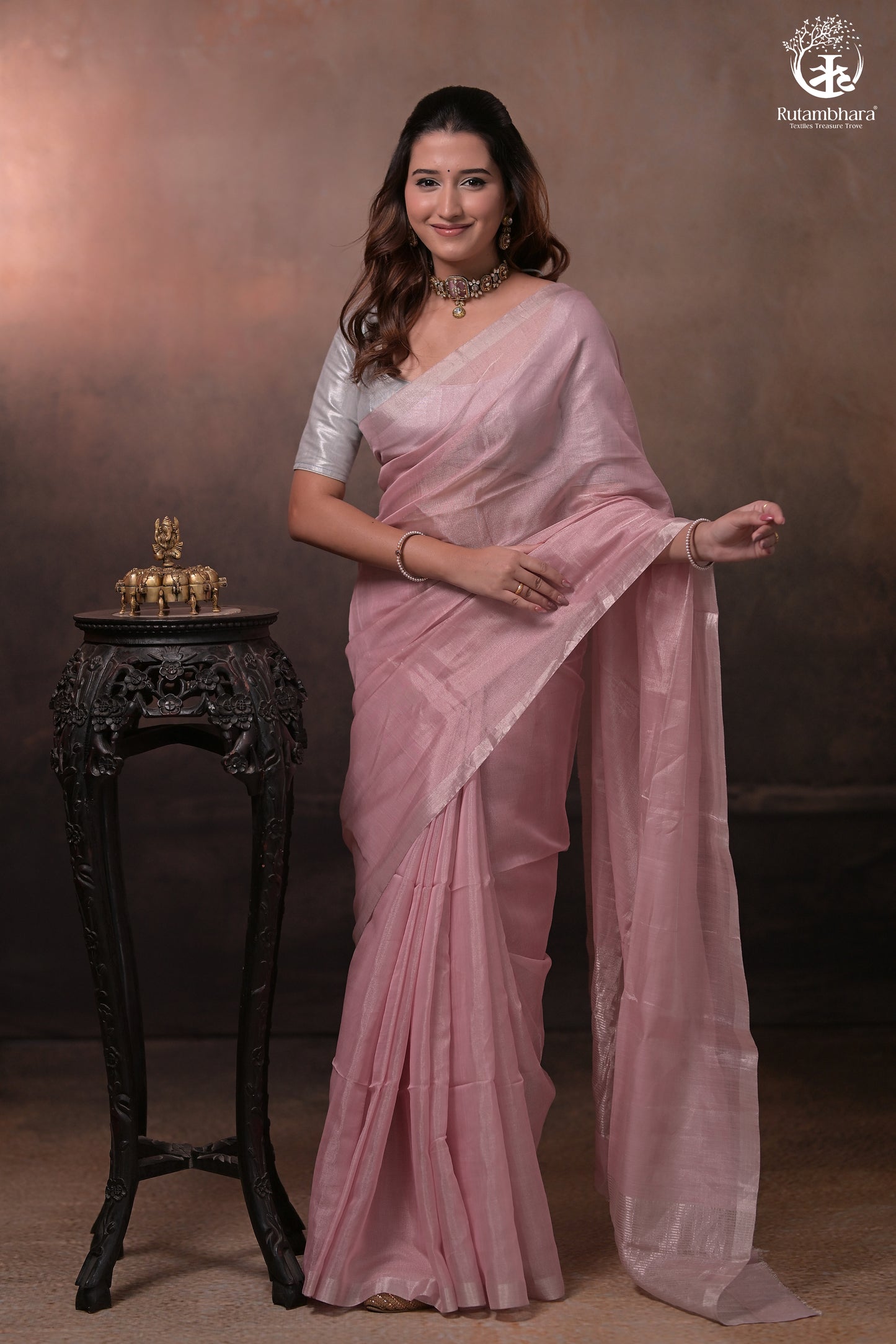 Woman in a light pink saree standing next to a small table with decorative items against a brown background.
