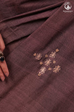 Close-up of a wine coloured saree  fabric with intricate mirror work  embroidery surrounding extra weft woven chomuk buta, featuring the brand 'Rutambhara' in the corner.